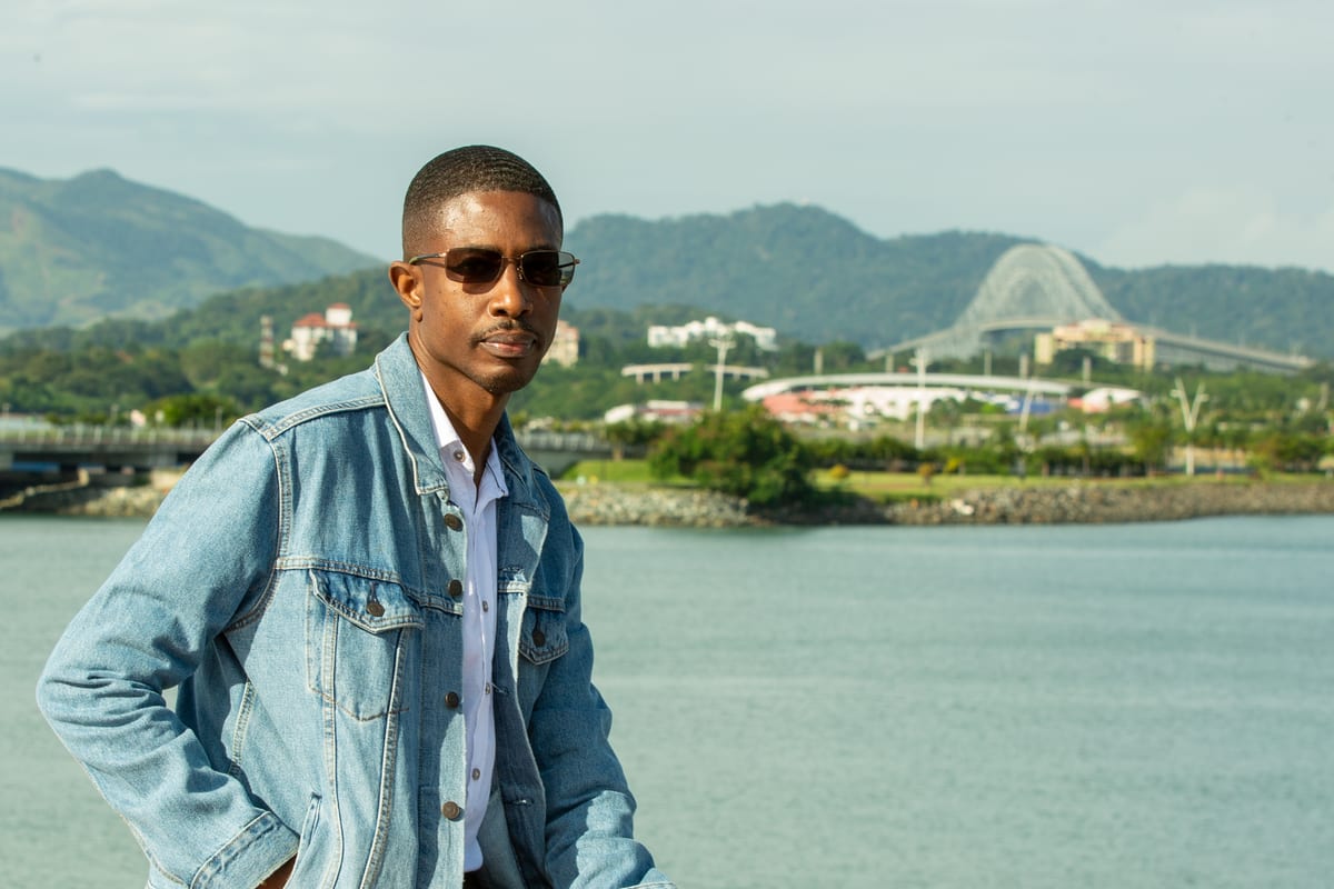 A man in a denim jacket and sunglasses with mountains and water behind him