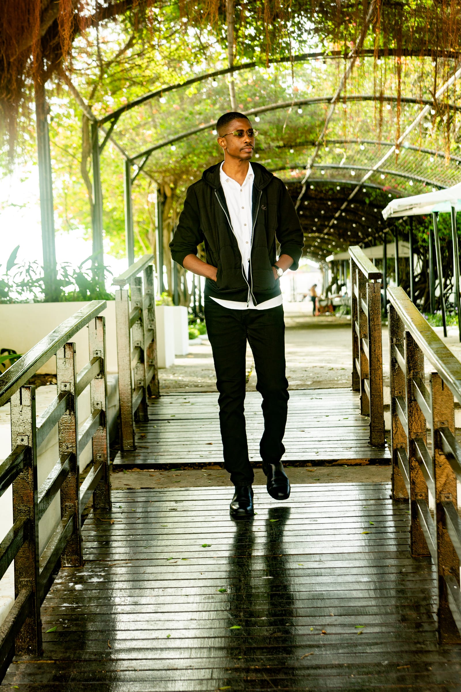 A man walking toward the camera along a trellised wooden boardwalk in a black hooded jacket and white shirt