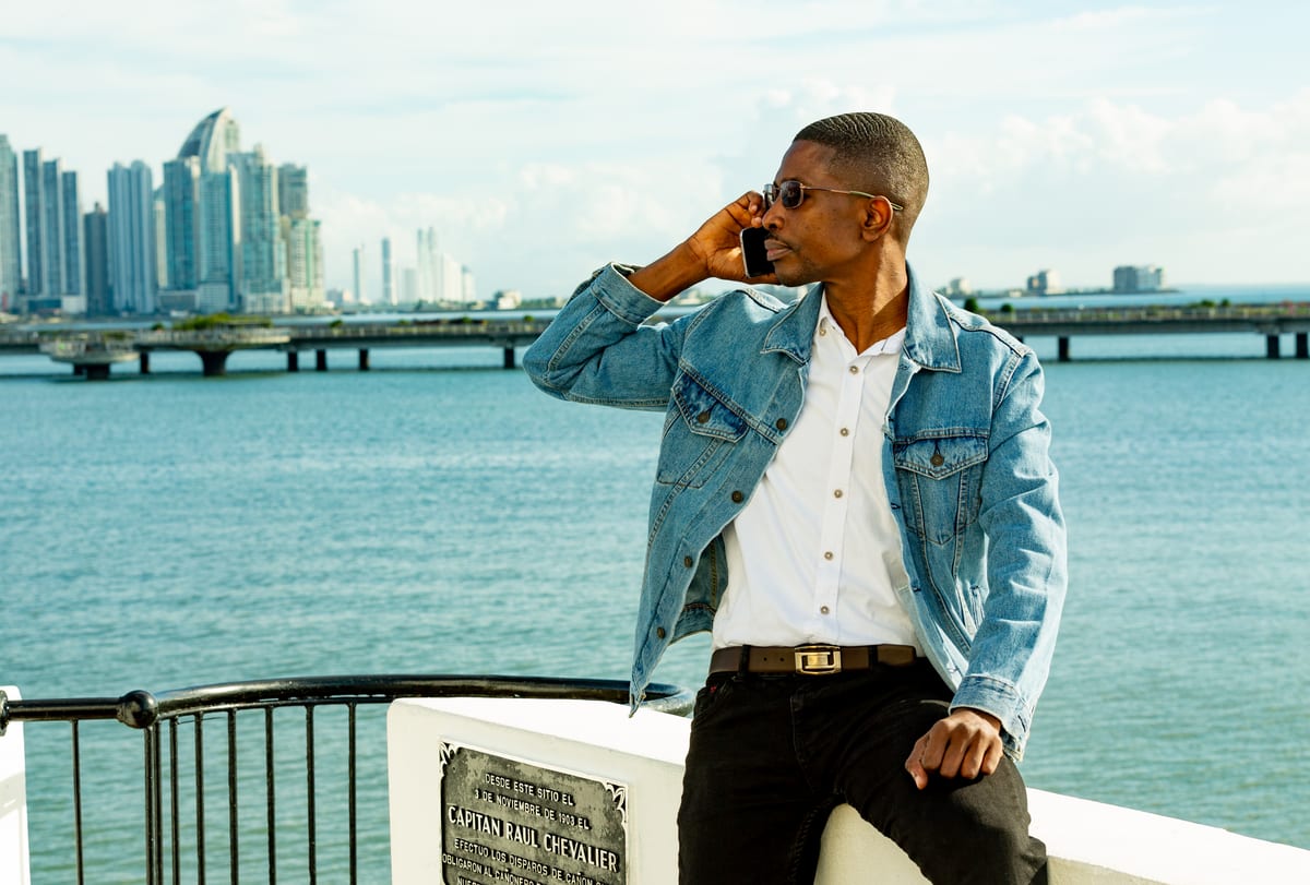 A man in a denim jacket on the phone by the waterfront with a city skyline behind