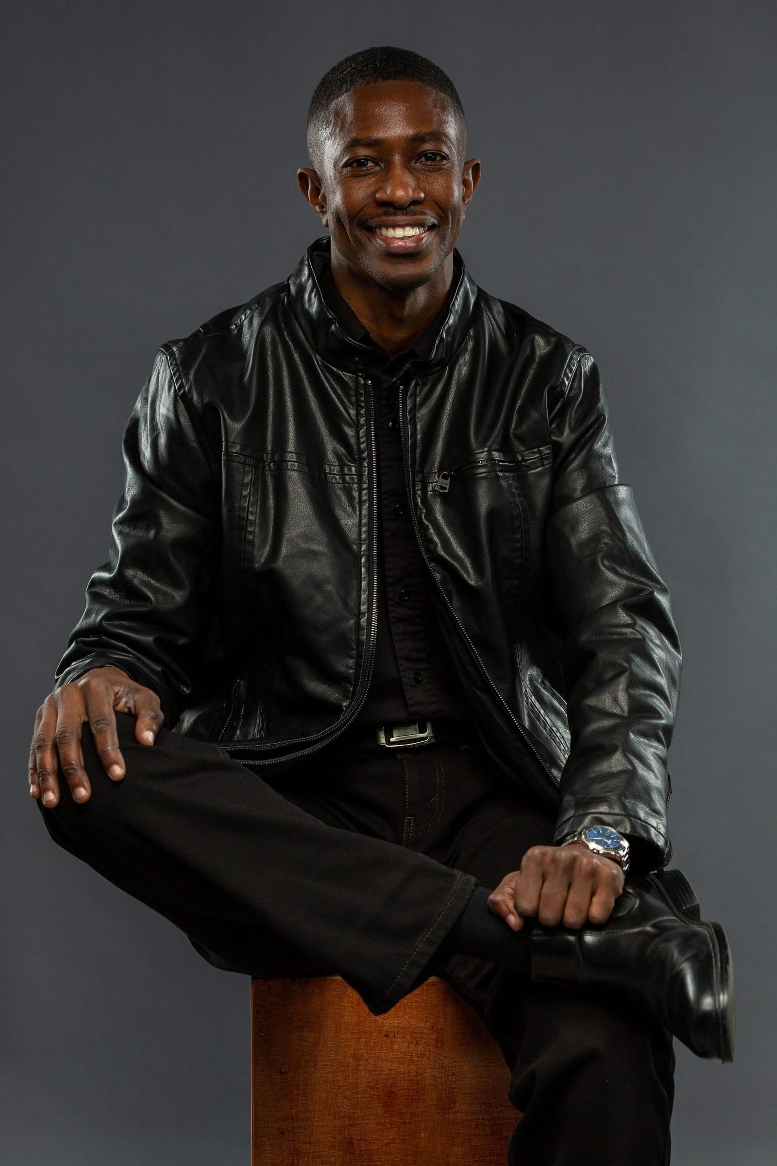 Studio portrait of a man seated in a black leather jacket, smiling