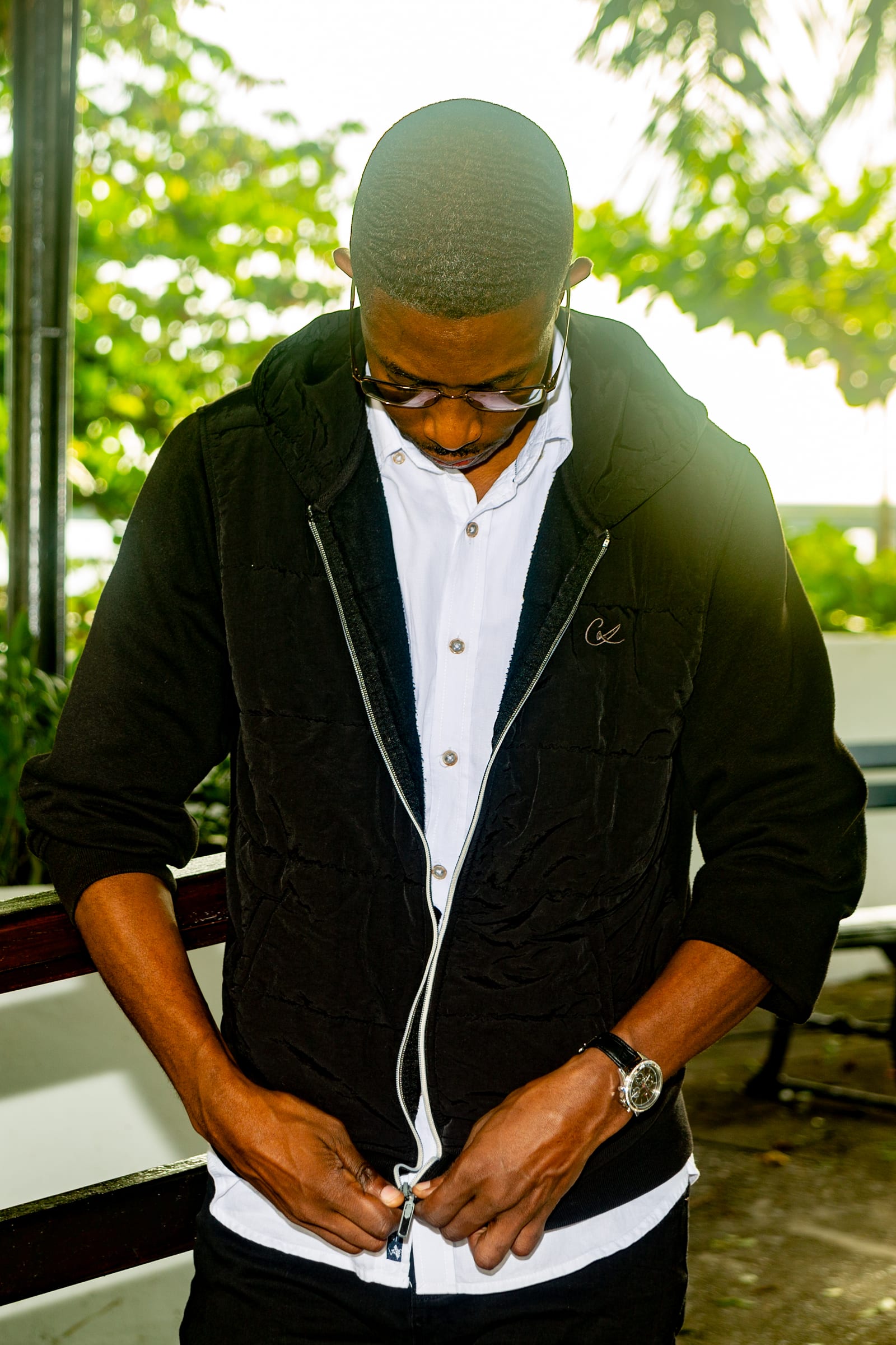 A man looking down as he adjusts his black jacket over a white shirt, soft greenery behind