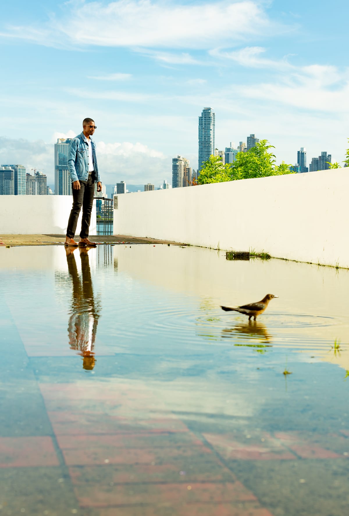 A man in a denim jacket standing on a rooftop against a city skyline, mirrored in a rain puddle