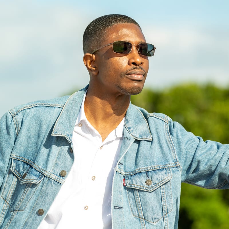 A smiling man in a denim jacket and white shirt under a bright summer sky