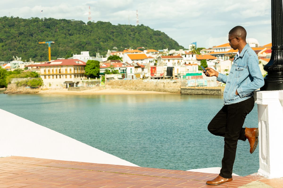 A man in a denim jacket leaning against a lamppost beside a colonial waterfront town